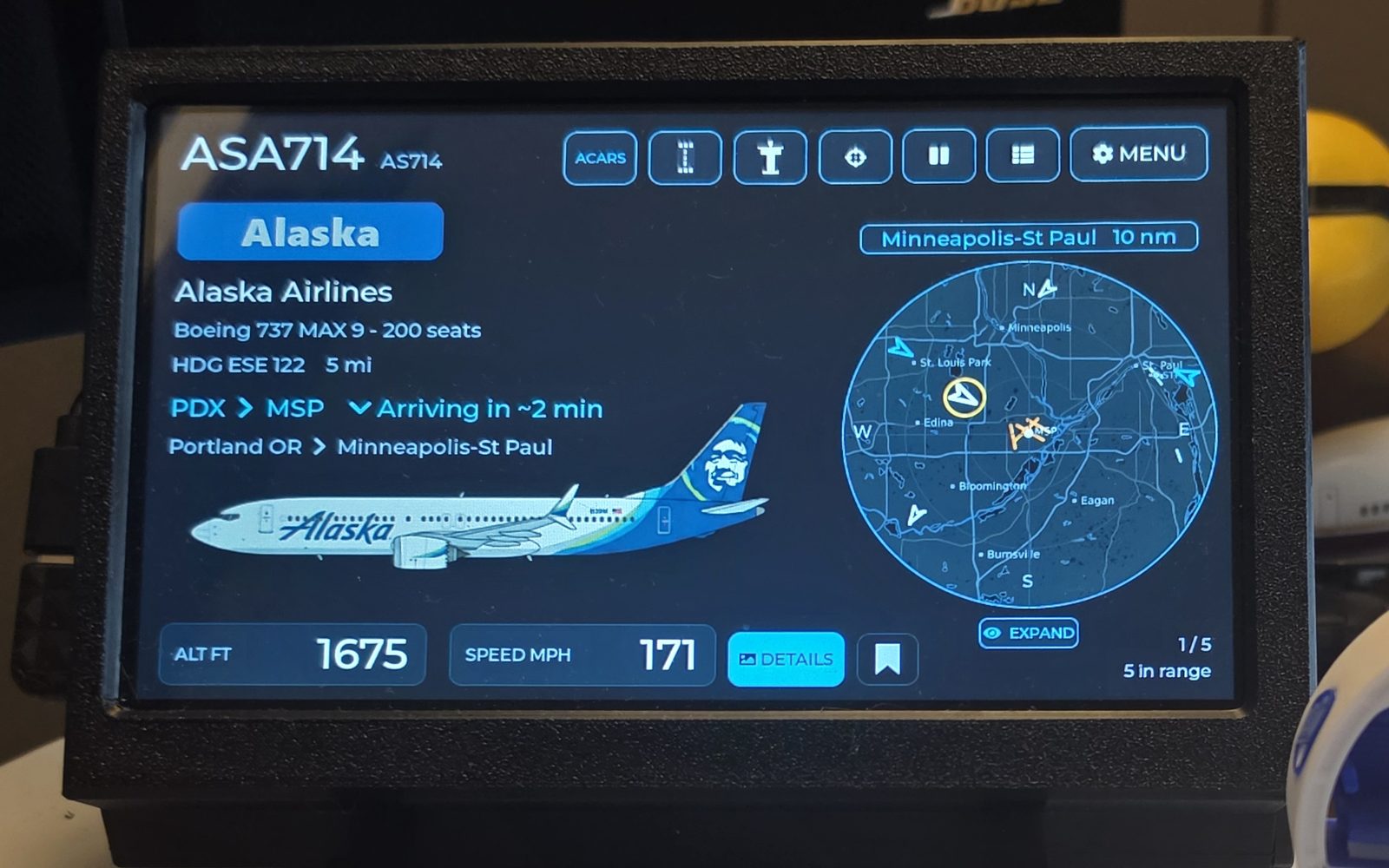 A WingsAbove 7-inch flight tracker on a desk, its screen showing flight ASA714 — a Boeing 737 MAX 9 from Portland, arriving Minneapolis-St Paul in about two minutes at 1,675 ft — beside a live radar map
