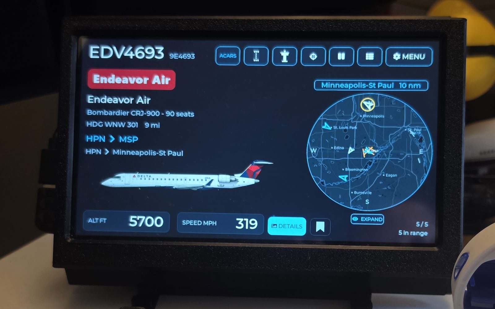 A WingsAbove 7-inch flight tracker on a desk showing flight EDV4693, a Bombardier CRJ-900 inbound to Minneapolis-St Paul at 5,700 ft and 319 mph, beside a live radar map